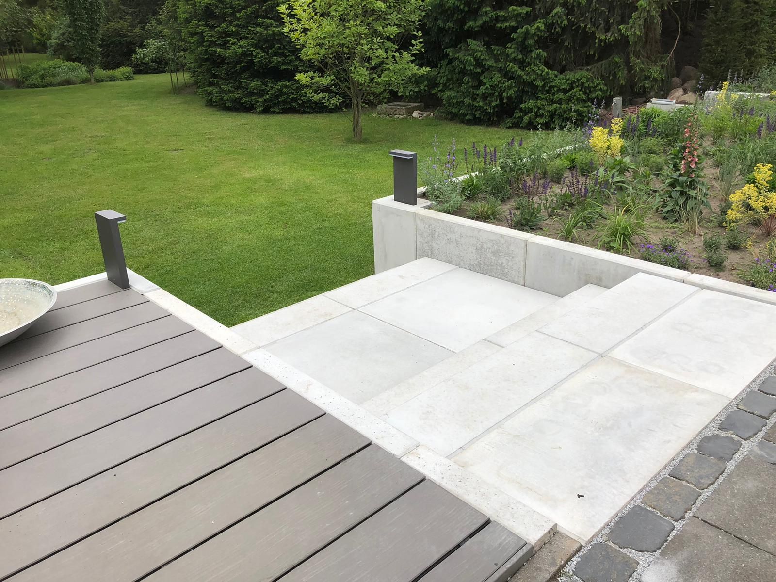 Eine Terrasse mit hellgrauen Steinstufen und dunklem Holzdeck führt zu einer Rasen- und Gartenfläche.