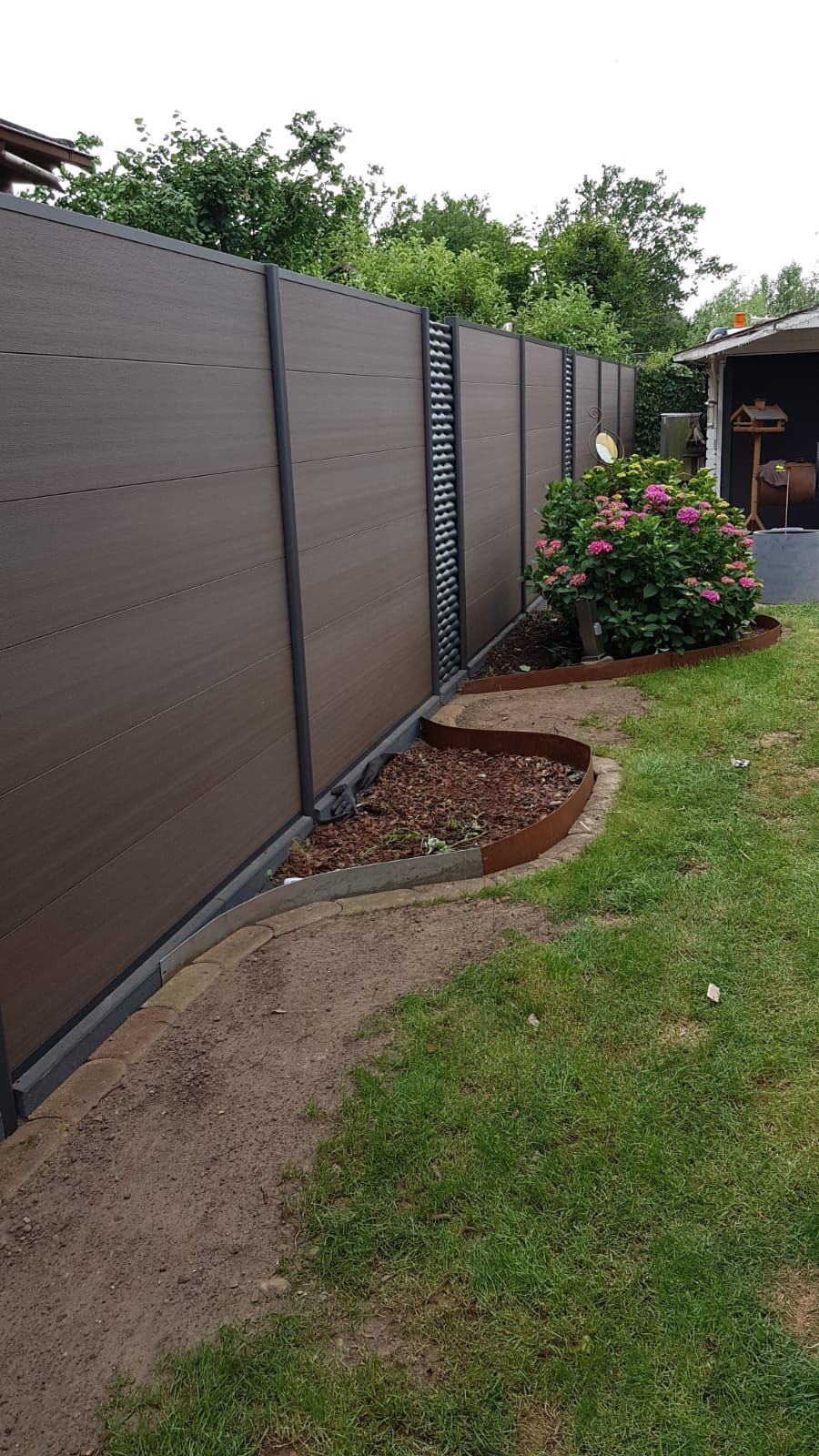 Brauner Sichtschutzzaun in einem grünen Garten mit einer Mulchschicht und blühenden Sträuchern.