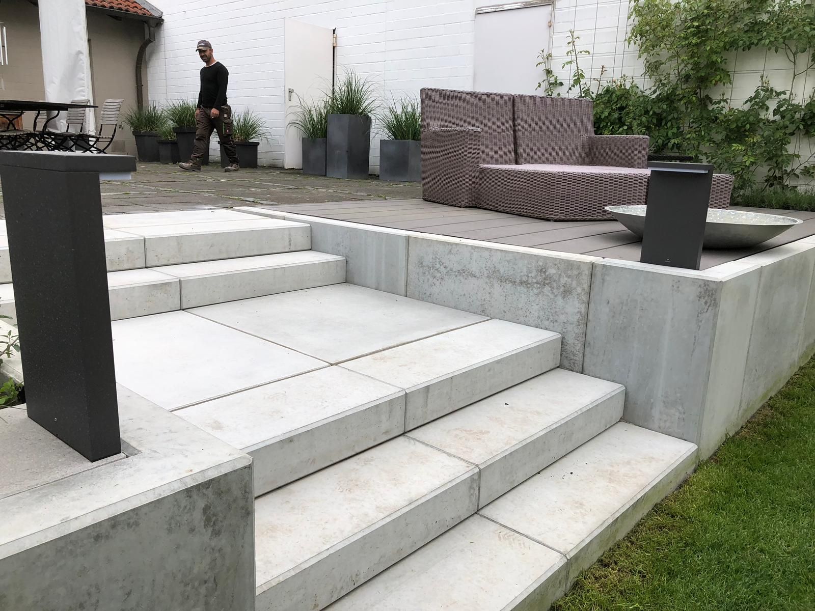 Über eine Außentreppe aus Beton gelangt man auf eine Terrasse mit Sitzgelegenheiten; im Hintergrund geht eine Person vorbei.