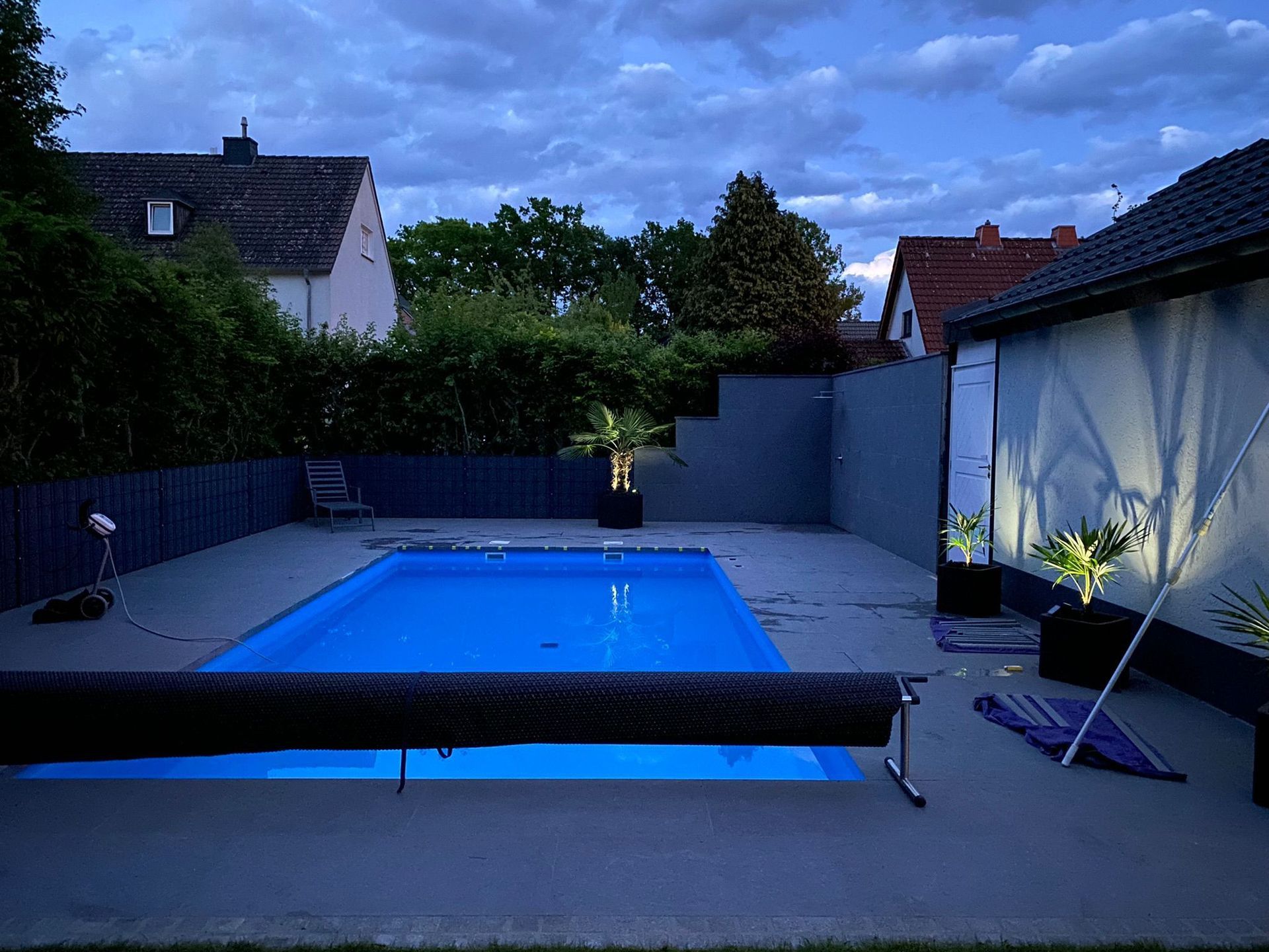 Pool mit blauem Wasser in der Abenddämmerung, umgeben von einer Betonterrasse, einem Zaun und Gebäuden.