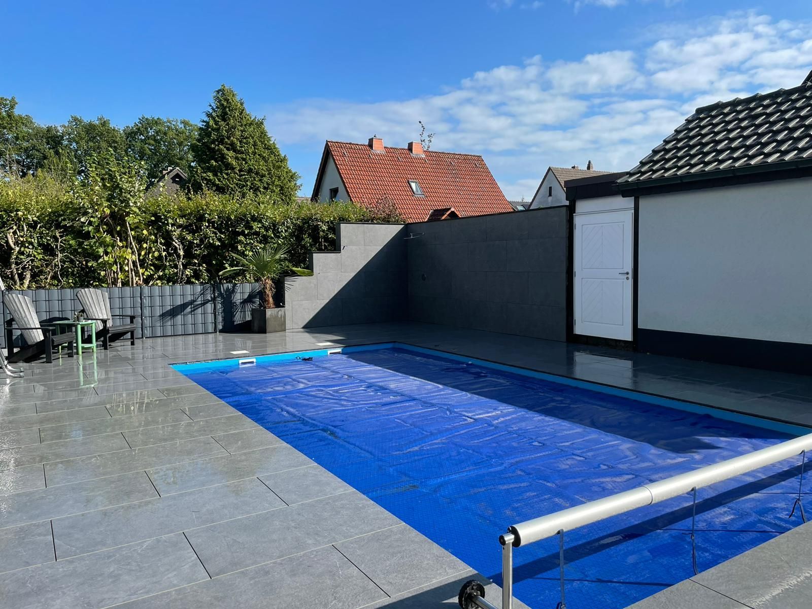 Ein blau überdachter rechteckiger Pool in einem gepflasterten Hof neben einer grauen Mauer und einem weißen Abstellgebäude.