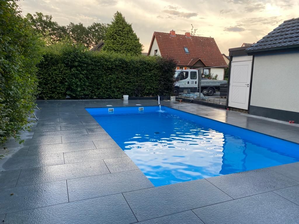 Rechteckiger blauer Pool, umgeben von einer dunkelgrau gefliesten Terrasse. Häuser und Bäume im Hintergrund. Bewölkter Himmel.