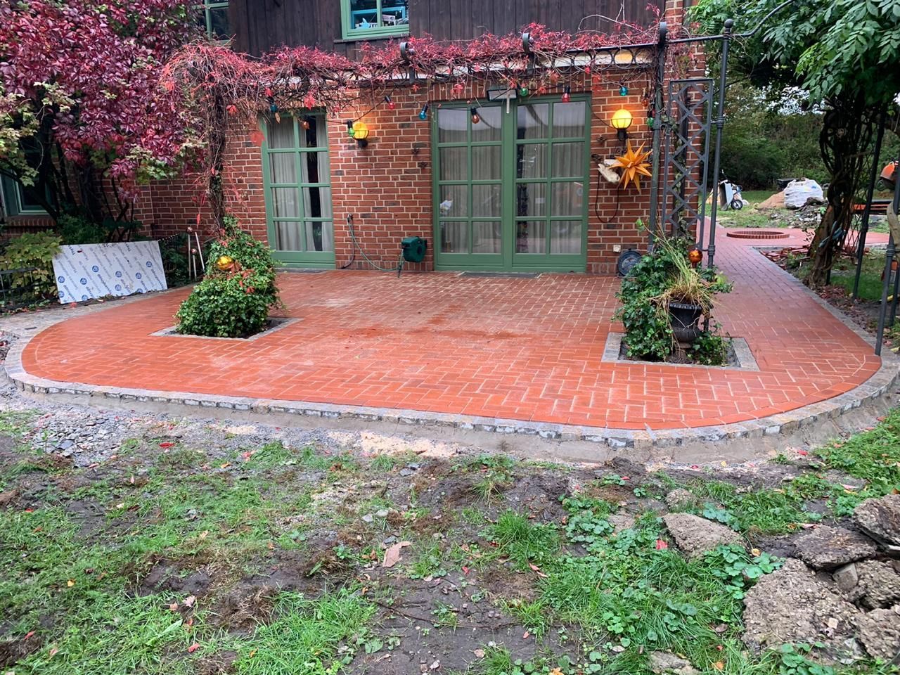 Terrasse aus roten Ziegelsteinen mit grüner Tür, umgeben von Grünpflanzen und einer Ziegelsteineinfassung.
