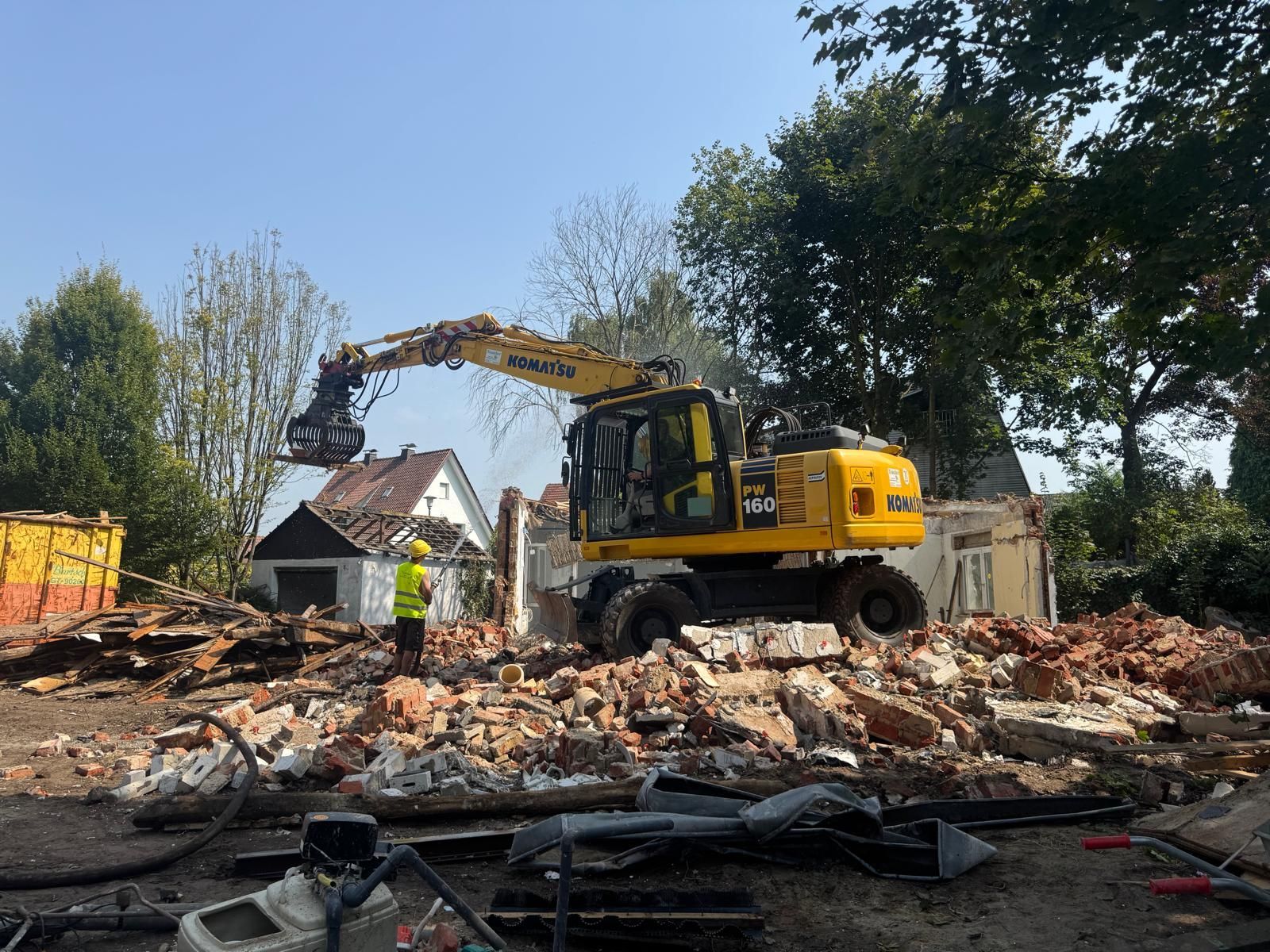 Ein gelber Bagger reißt ein Gebäude ab; ein Arbeiter in einer Sicherheitsweste steht daneben.