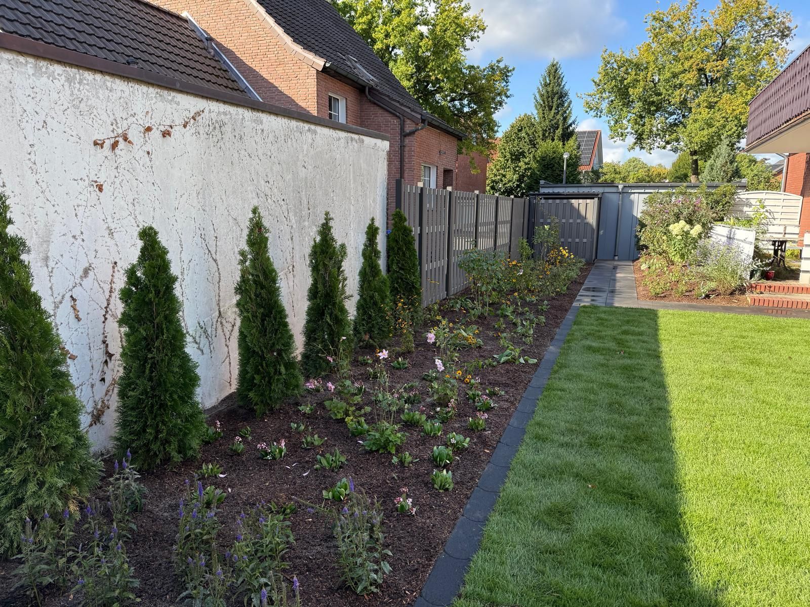 Grüne Sträucher, Blumen und ein frisch gemähter Rasen säumen eine weiße Mauer und einen Holzzaun in einem Garten.