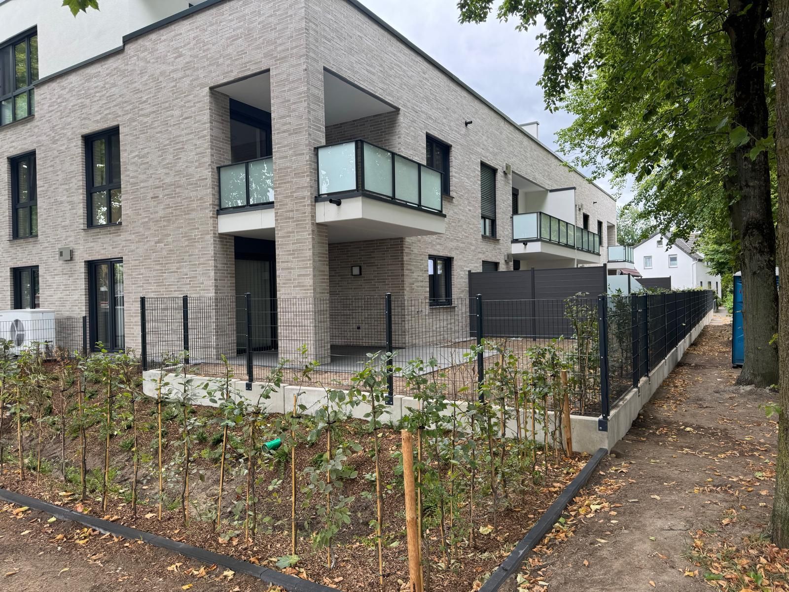 Modernes Backstein-Mehrfamilienhaus mit Balkonen, schwarzem Zaun und kleinen Bäumen entlang des Gehwegs.