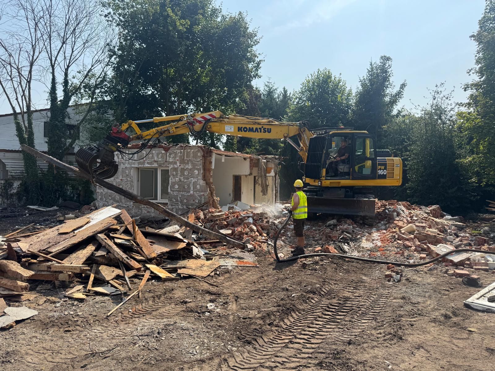 Ein gelber Bagger reißt ein Backsteingebäude ab, ein Arbeiter besprüht es mit Wasser. Draußen liegen Trümmer verstreut.