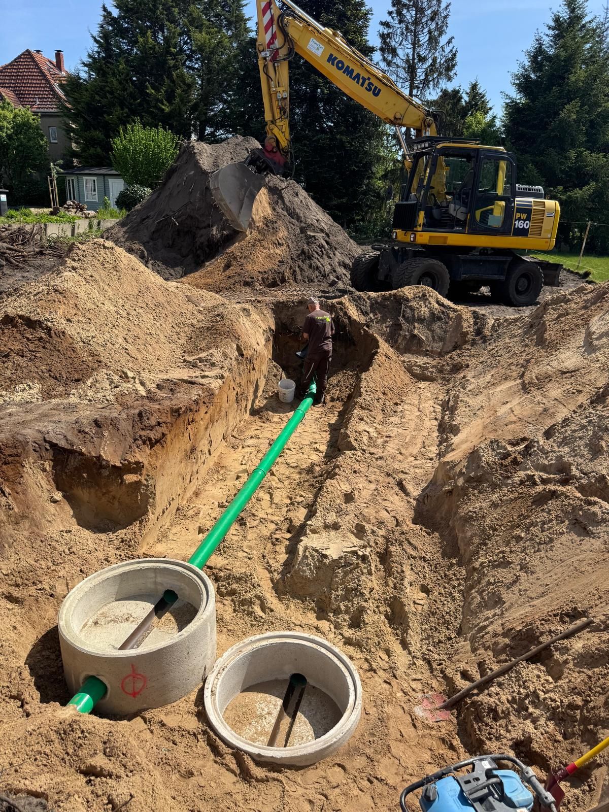 Ein Bagger hebt einen Graben aus und installiert im Freien in sandigem Boden ein grünes Rohrsystem und Klärgruben aus Beton.