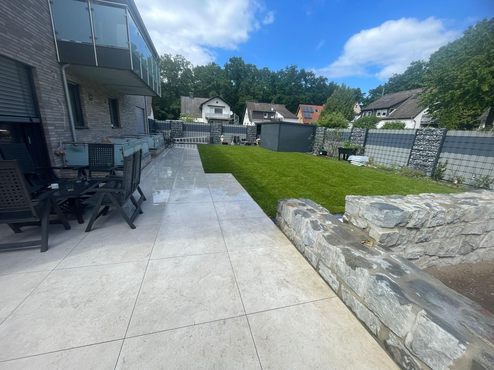 Terrasse mit Steinmauer, Stühlen und Rasenfläche. Gebäude und Bäume im Hintergrund unter blauem Himmel.