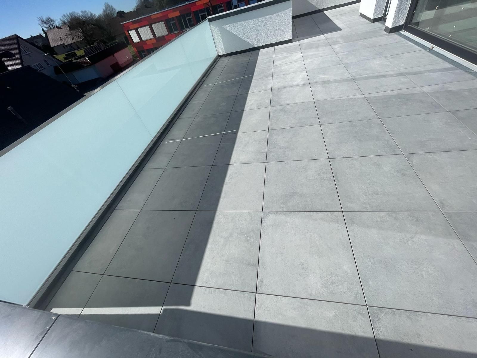 Außenansicht der Dachterrasse mit grauen Fliesen, Glasgeländer und einem Gebäude im Hintergrund.