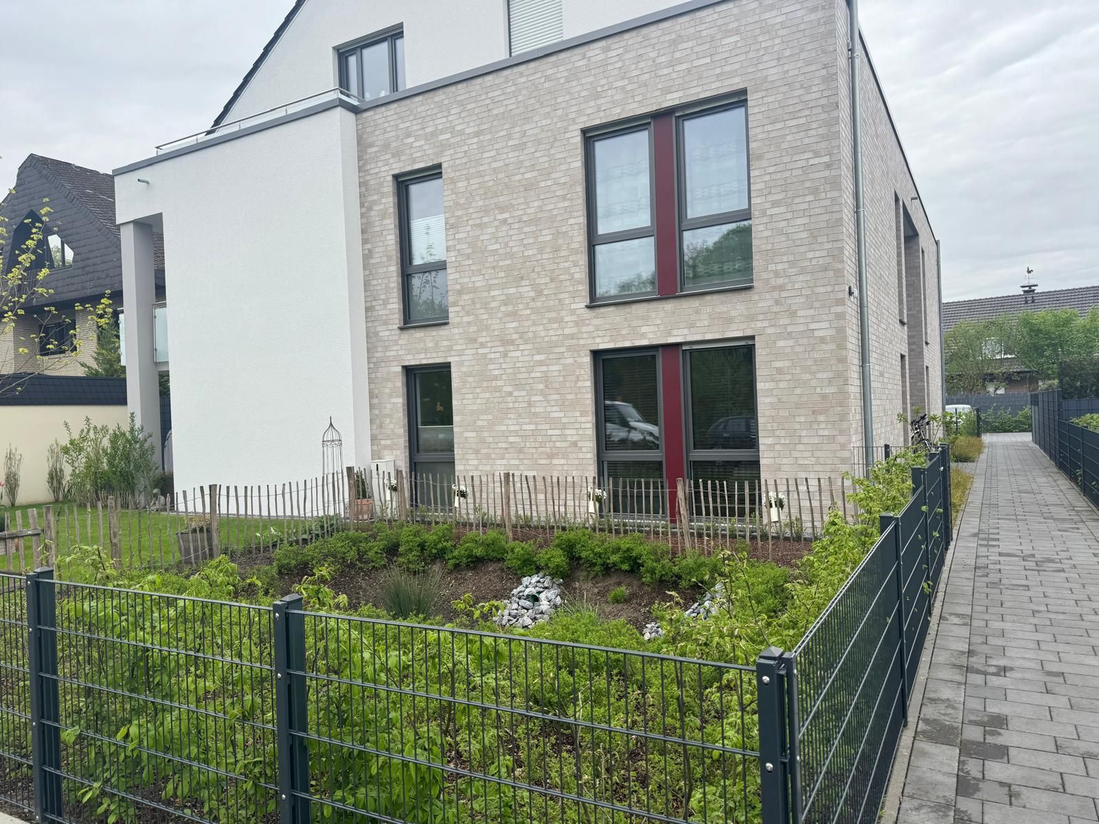 Modernes Backsteingebäude mit großen Fenstern und einem kleinen Garten hinter einem schwarzen Zaun.