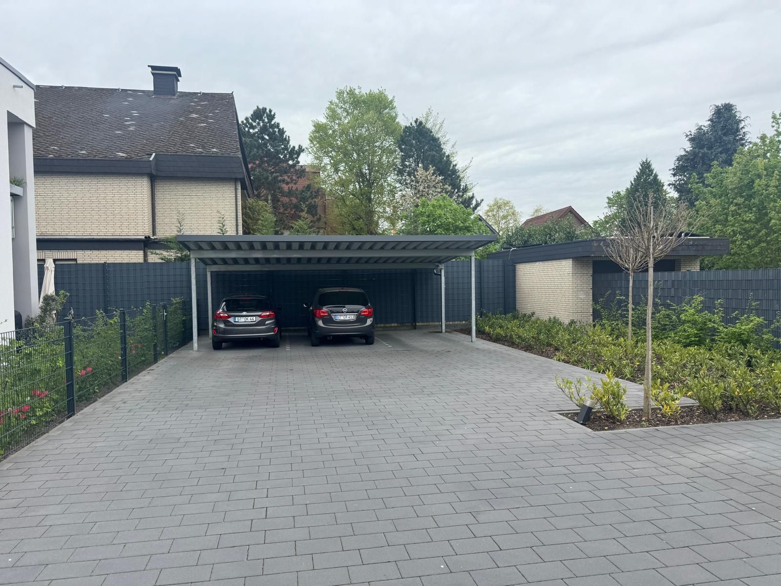 Zwei Autos parken unter einem Carport auf einer gepflasterten Auffahrt; im Hintergrund sind ein Haus und Bäume zu sehen.