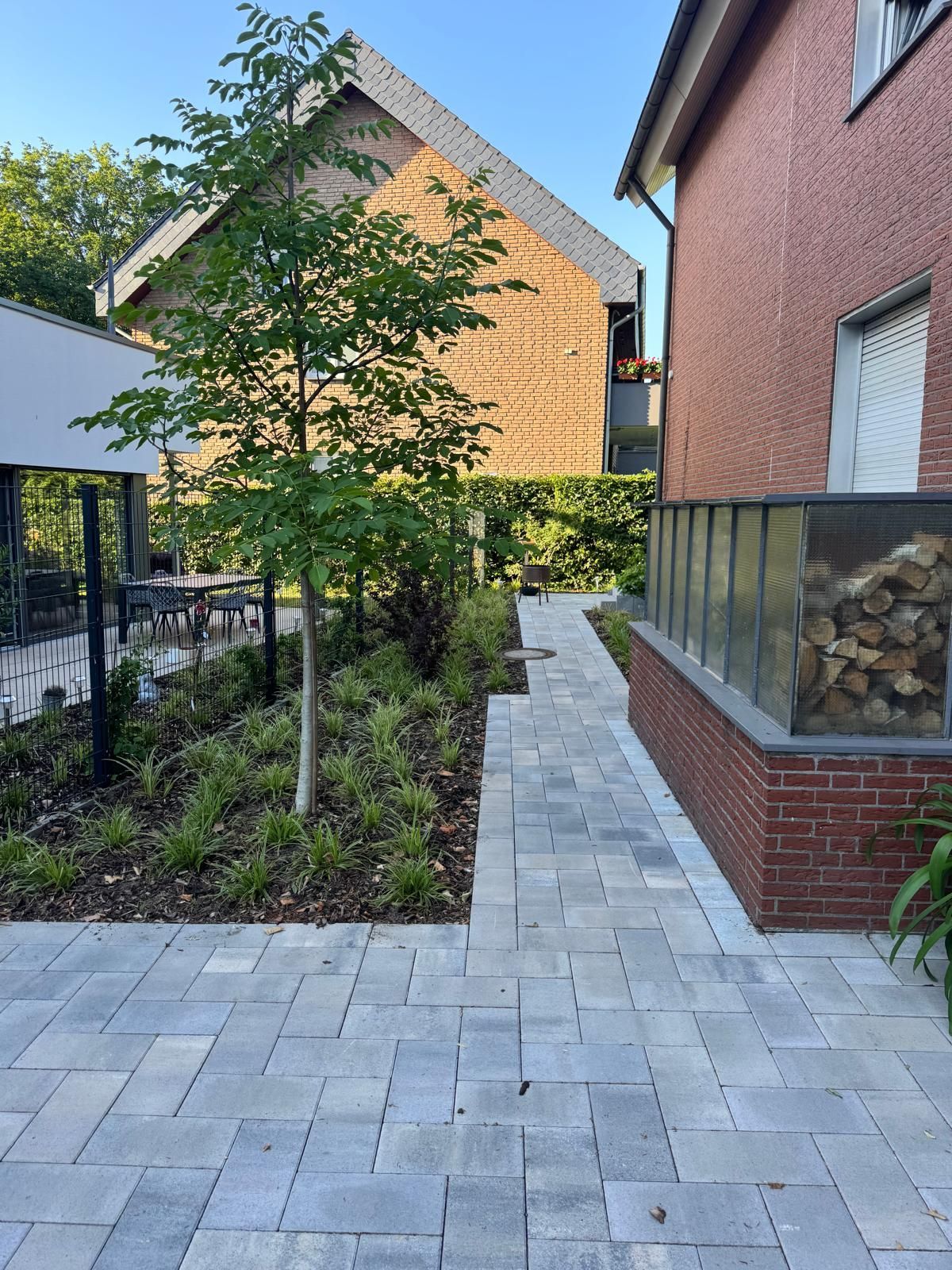 Ein Ziegelweg entlang einer Ziegelmauer mit Brennholzvorrat führt zu einem grün bepflanzten Garten und dem Haus.