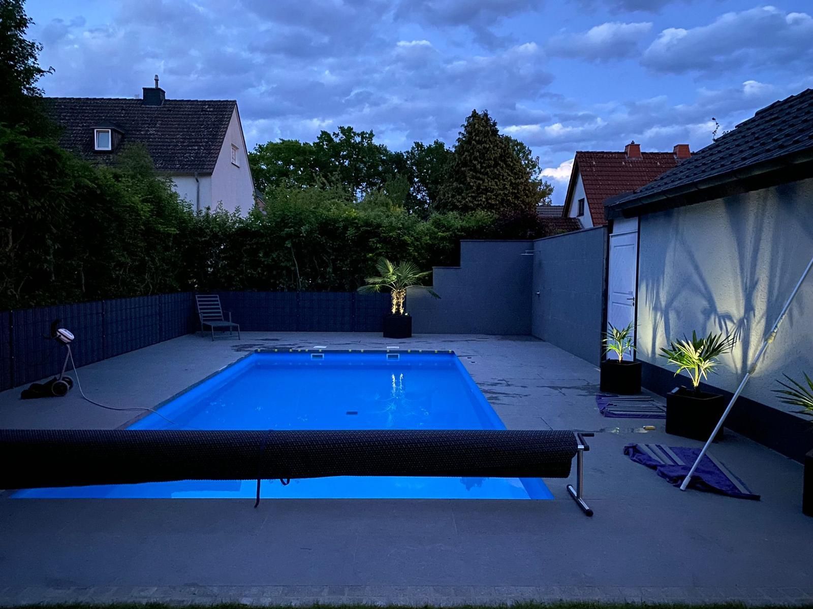 Hinterhofpool in der Abenddämmerung, blaues Wasser, dunkler Zaun, bewölkter Himmel, beleuchtet von Lampen.