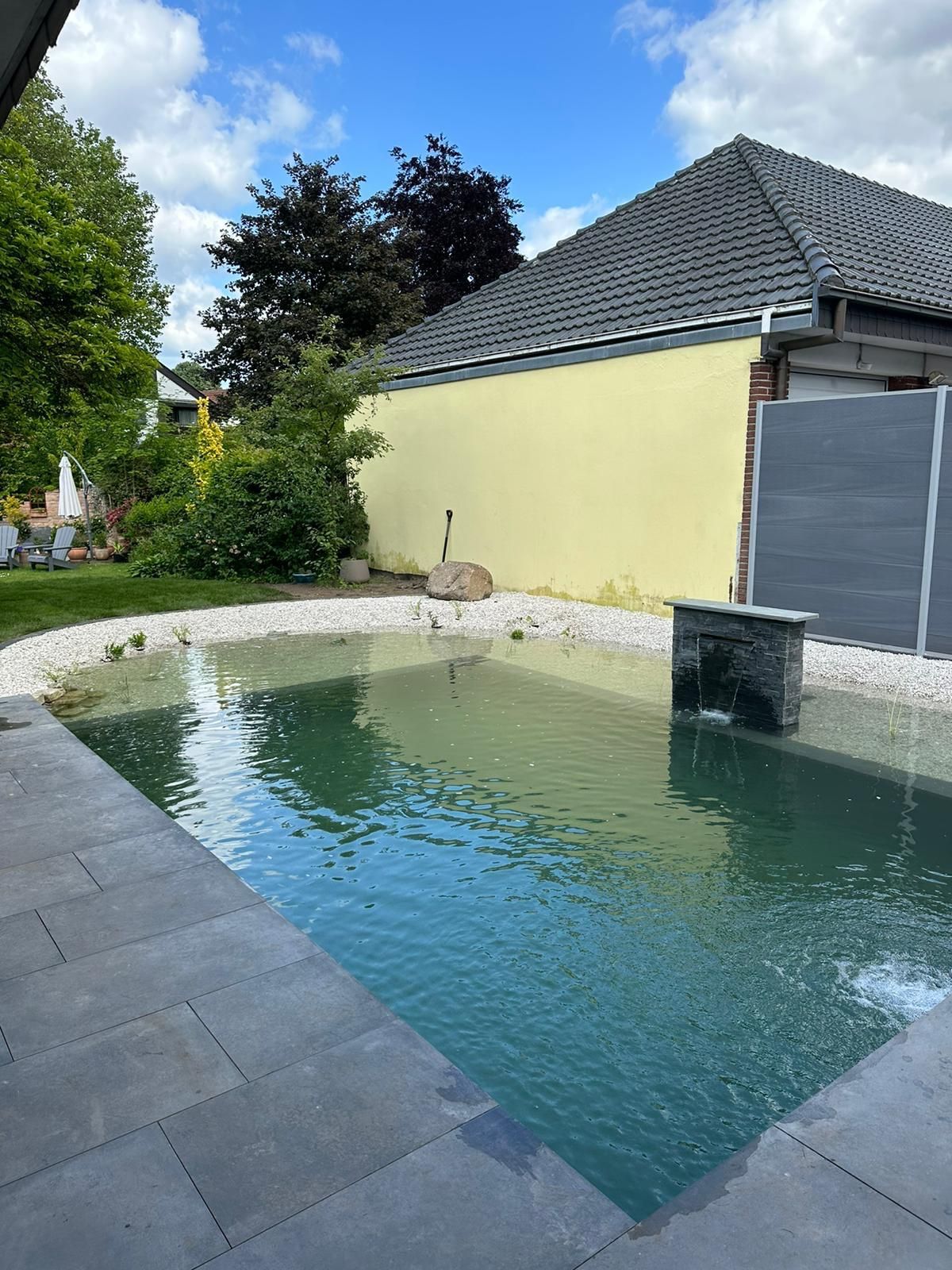 Teich in einem Hinterhof mit gelber Mauer, grauen Fliesen und weißen Steinen rund um den Teich.