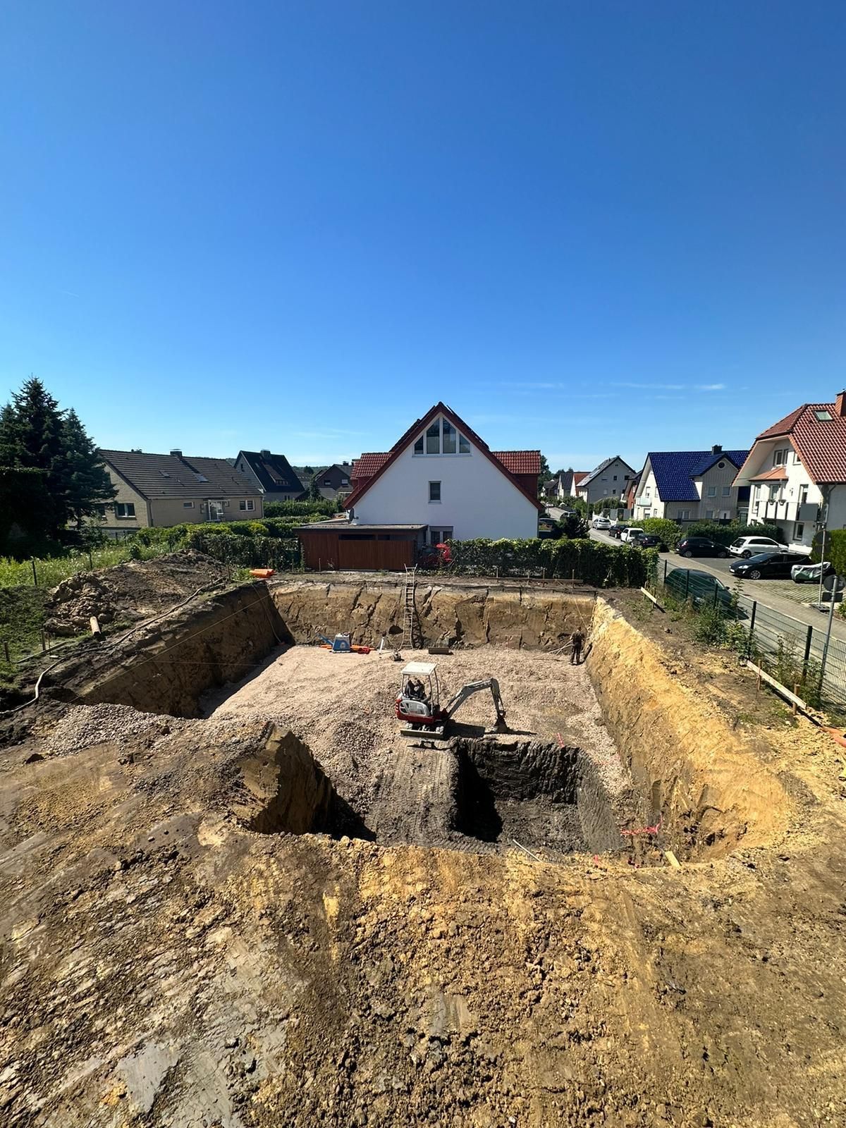 Baustelle: Ausgehobene Baugrube mit Bagger, im Hintergrund Wohnhäuser unter blauem Himmel.