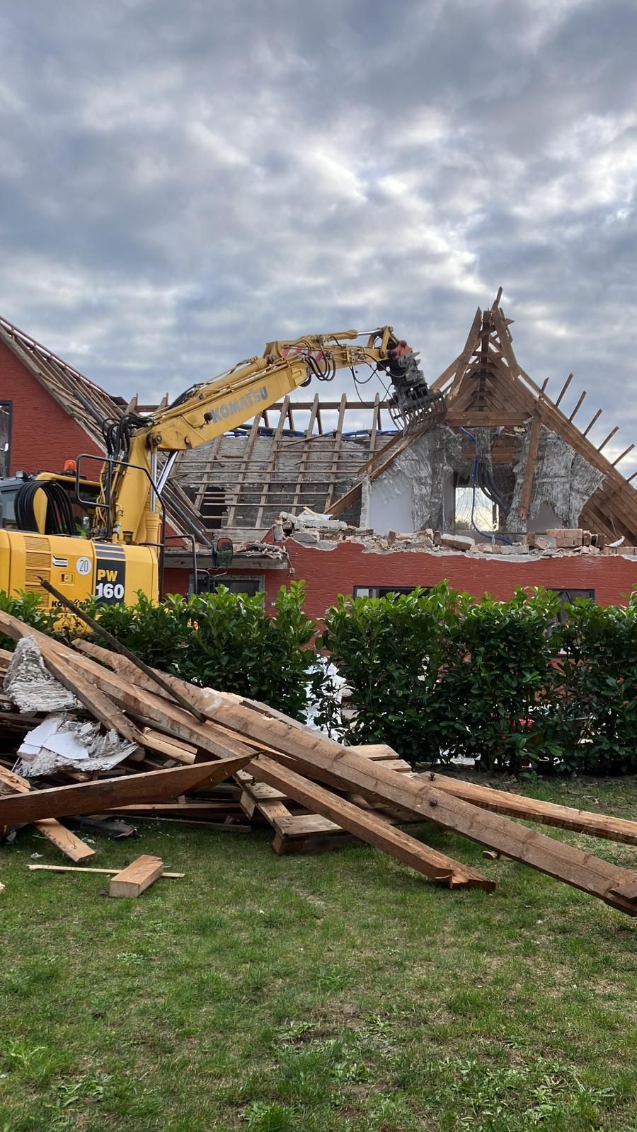 An einem bewölkten Tag reißt ein gelber Bagger ein Gebäude mit roter Backsteinfassade ab. Trümmer und Holzreste liegen auf dem Rasen.