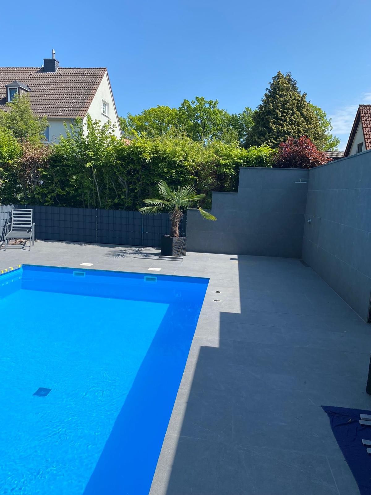 Schwimmbecken mit blauem Wasser und grauer Betonterrasse; im Hintergrund ein Sichtschutzzaun und Laubwerk.