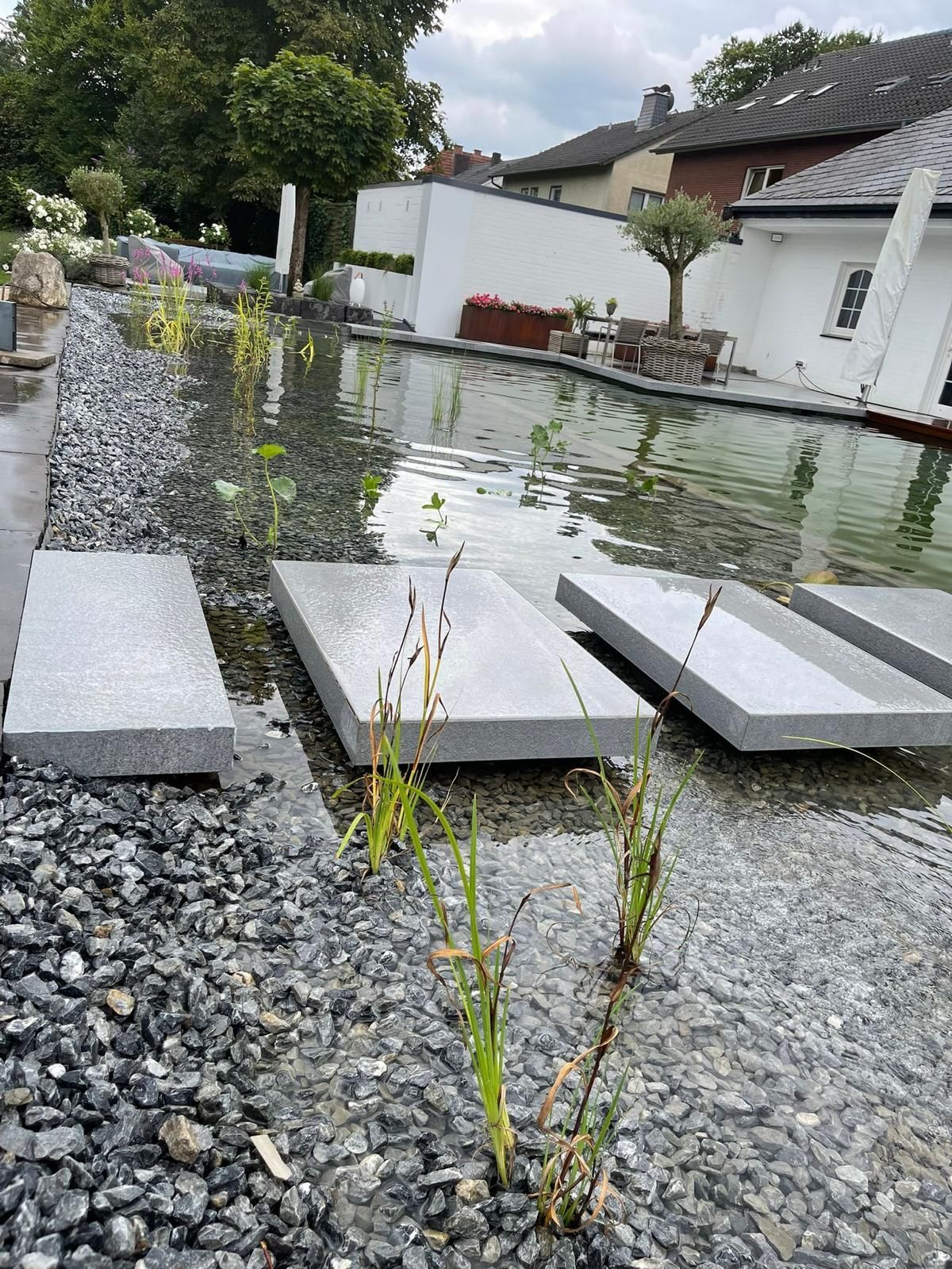 Steinplatten über einem Wasserspiel mit Kiesrändern, Pflanzen und einem weißen Gebäude im Hintergrund.