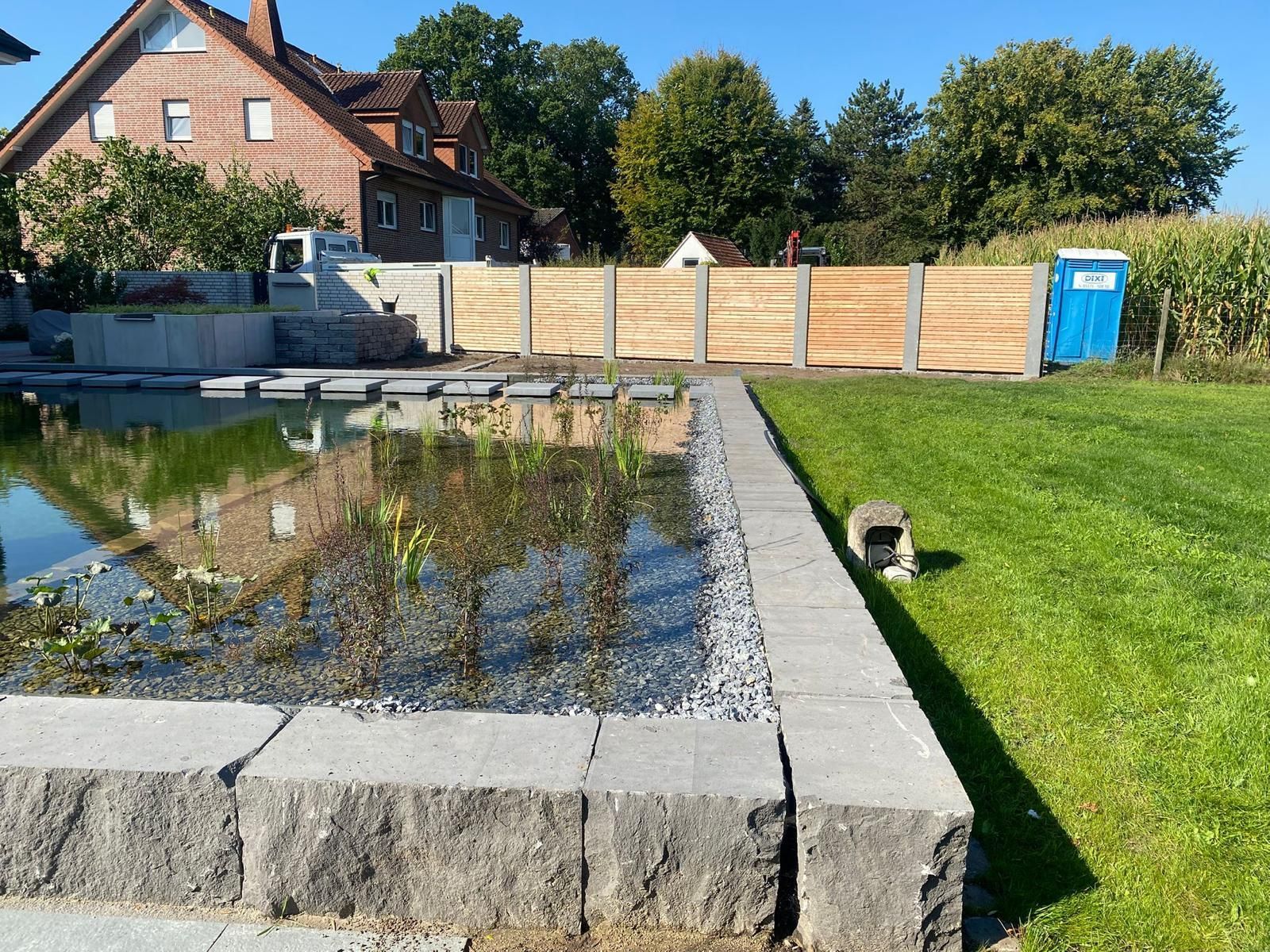 Teich mit Steineinfassung, Zaun und Rasen. Im Hintergrund ist ein Haus an einem sonnigen Tag zu sehen.