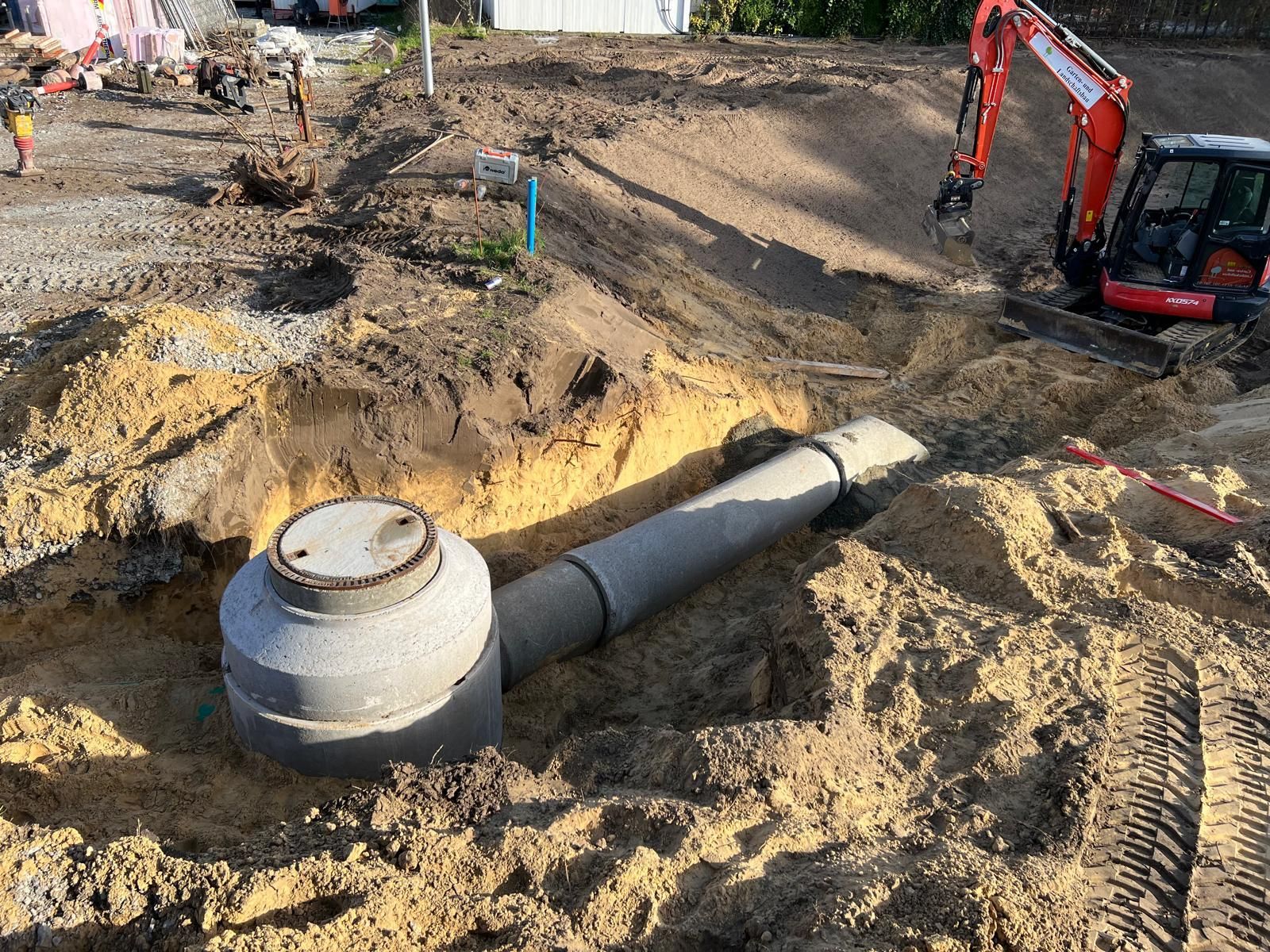 Aushubstelle mit Betontank und Rohren; im Hintergrund ein kleiner Bagger und Arbeiter.