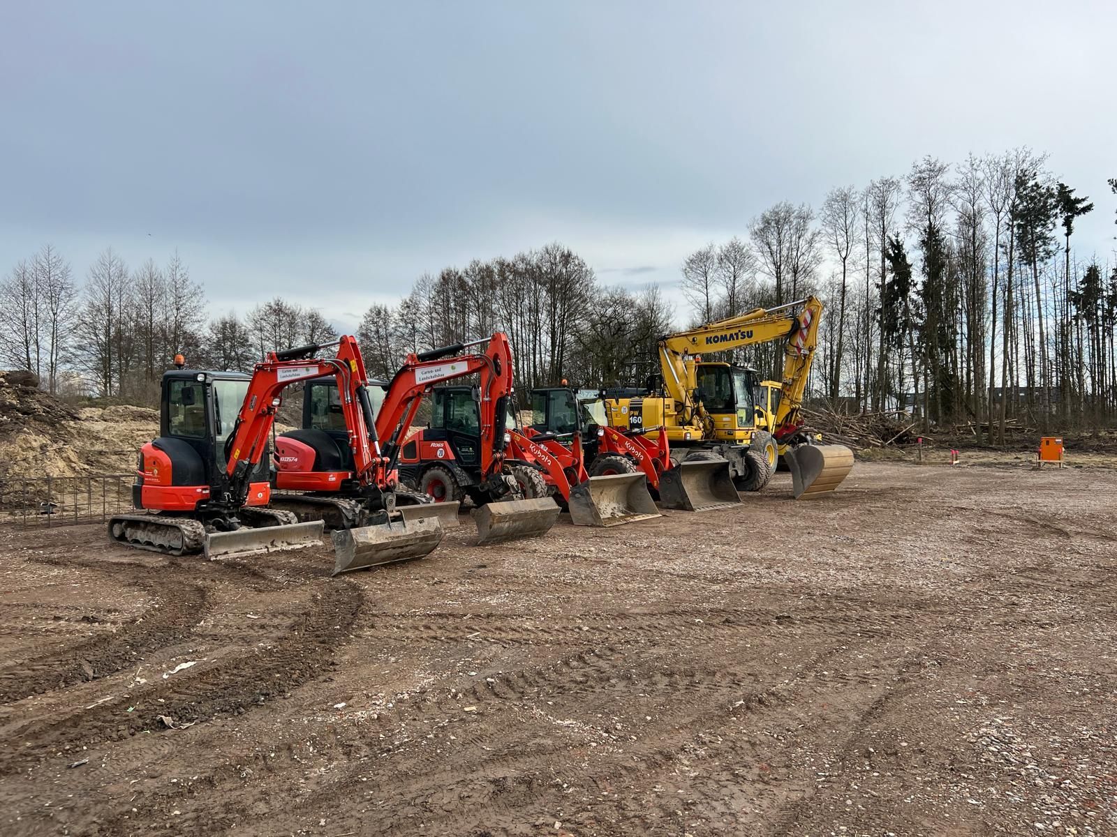Fünf Bagger stehen auf einer Baustelle. Rote und gelbe Baumaschinen vor bewölktem Himmel.