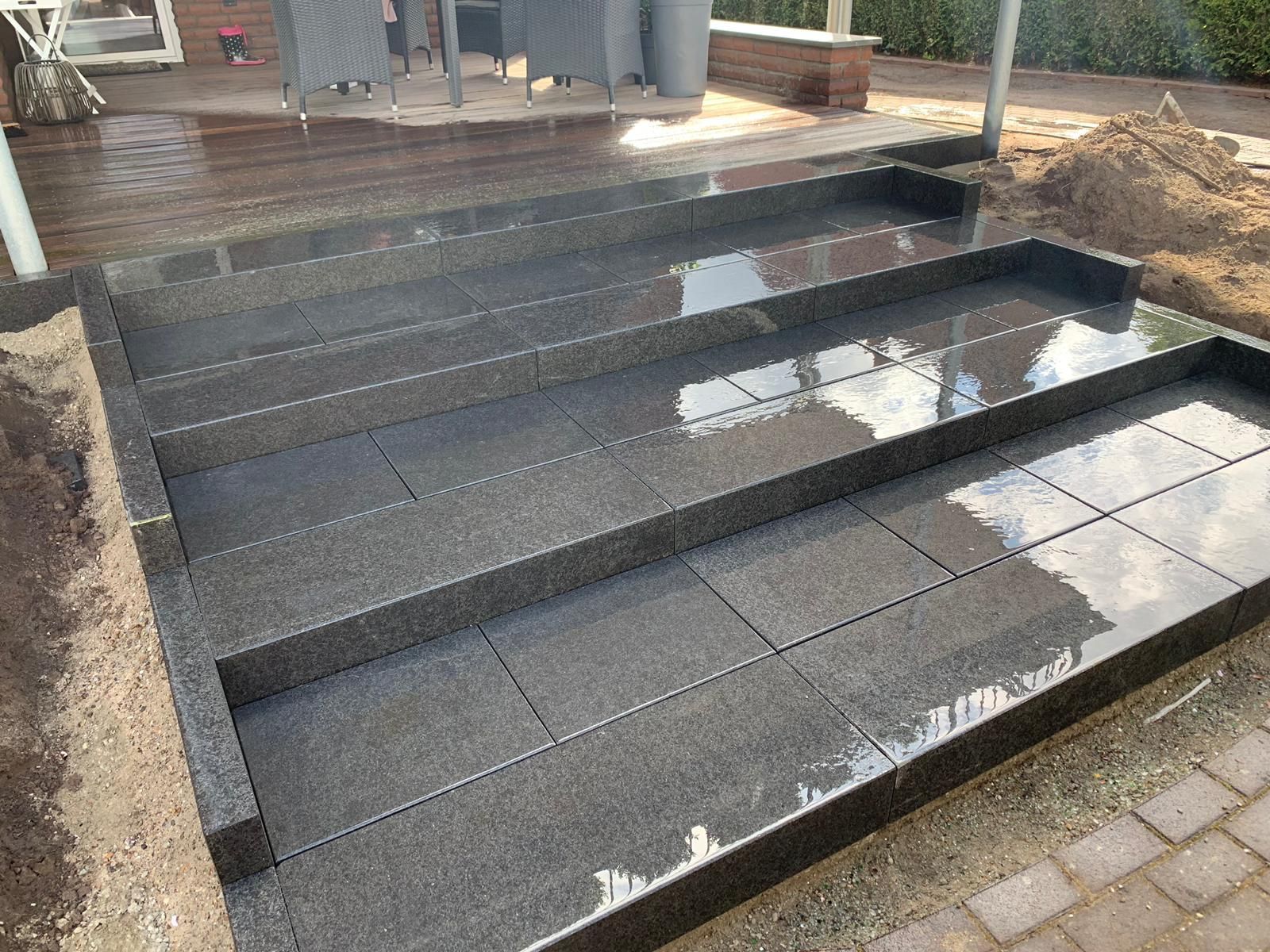 Neu errichtete Außentreppe aus dunkelgrauen Fliesen; nasse Oberfläche; angrenzend an unbefestigten Boden und eine Holzterrasse.