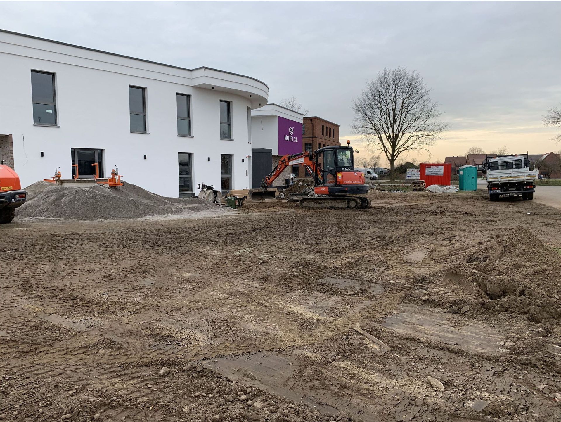 Baustelle mit einem weißen Gebäude, einem Bagger und Erdhaufen.