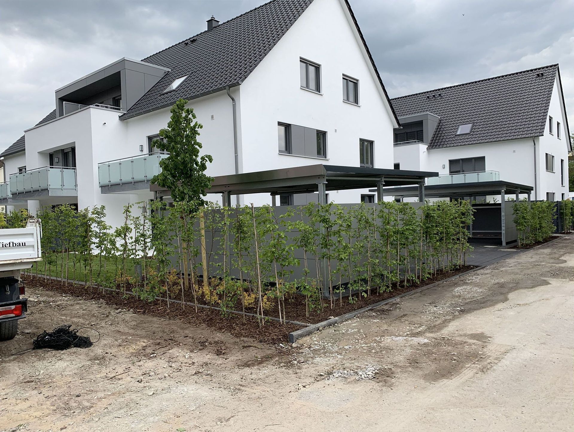 Moderne weiße Häuser mit grauen Dächern, Carports und einer neu gepflanzten grünen Hecke.