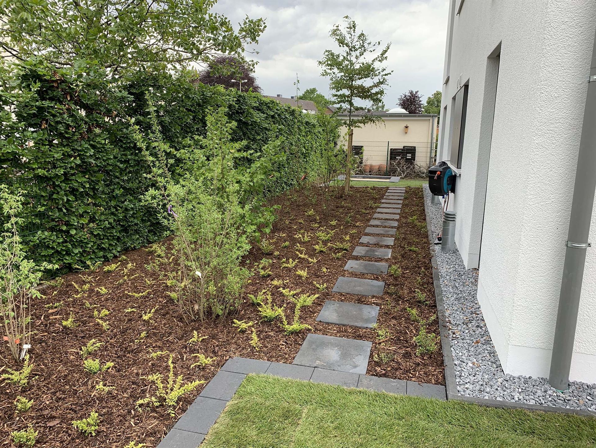 Ein Steinweg führt durch einen mit Mulch bedeckten und von Pflanzen gesäumten Garten, neben einem Gebäude.