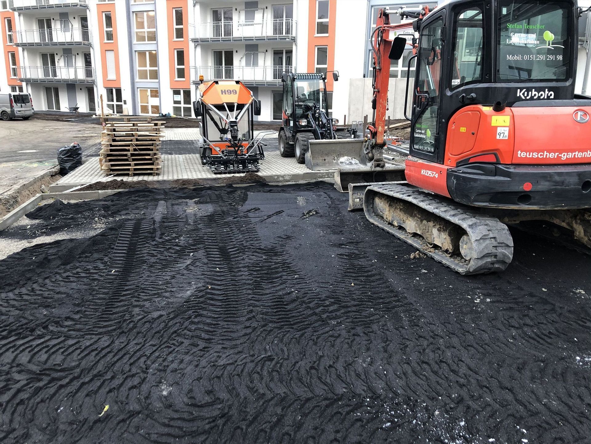 Baustelle mit Asphalt, Minibagger und anderen Baumaschinen. Im Hintergrund ein neues Gebäude.
