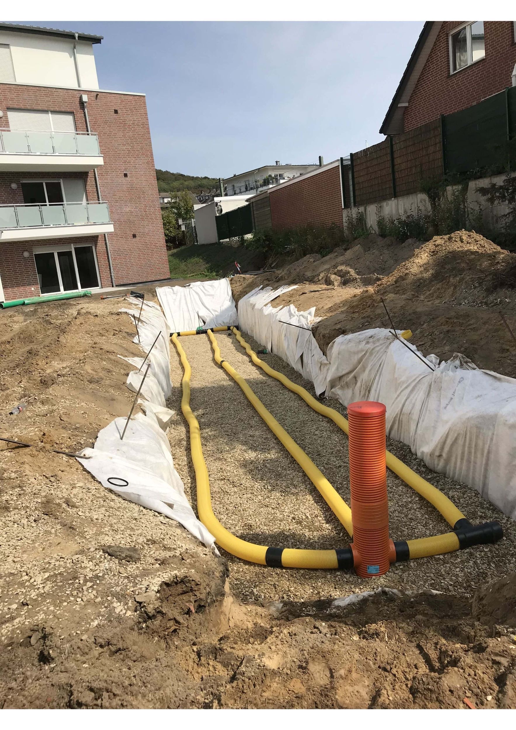 Baustelle mit gelben Rohren und Kies in einem Graben, neben einem Gebäude und einem Hügel.