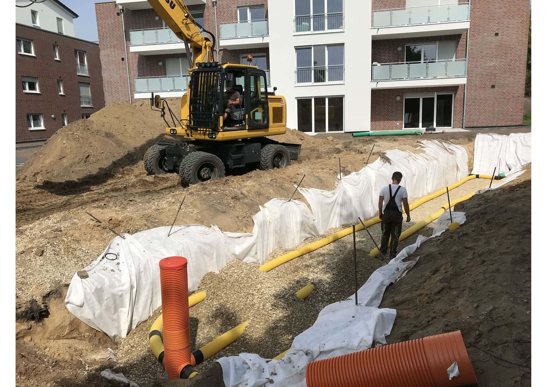 Baustelle mit Bagger, Arbeiter, Rohren und Sedimentbarrieren neben einem mehrstöckigen Gebäude.