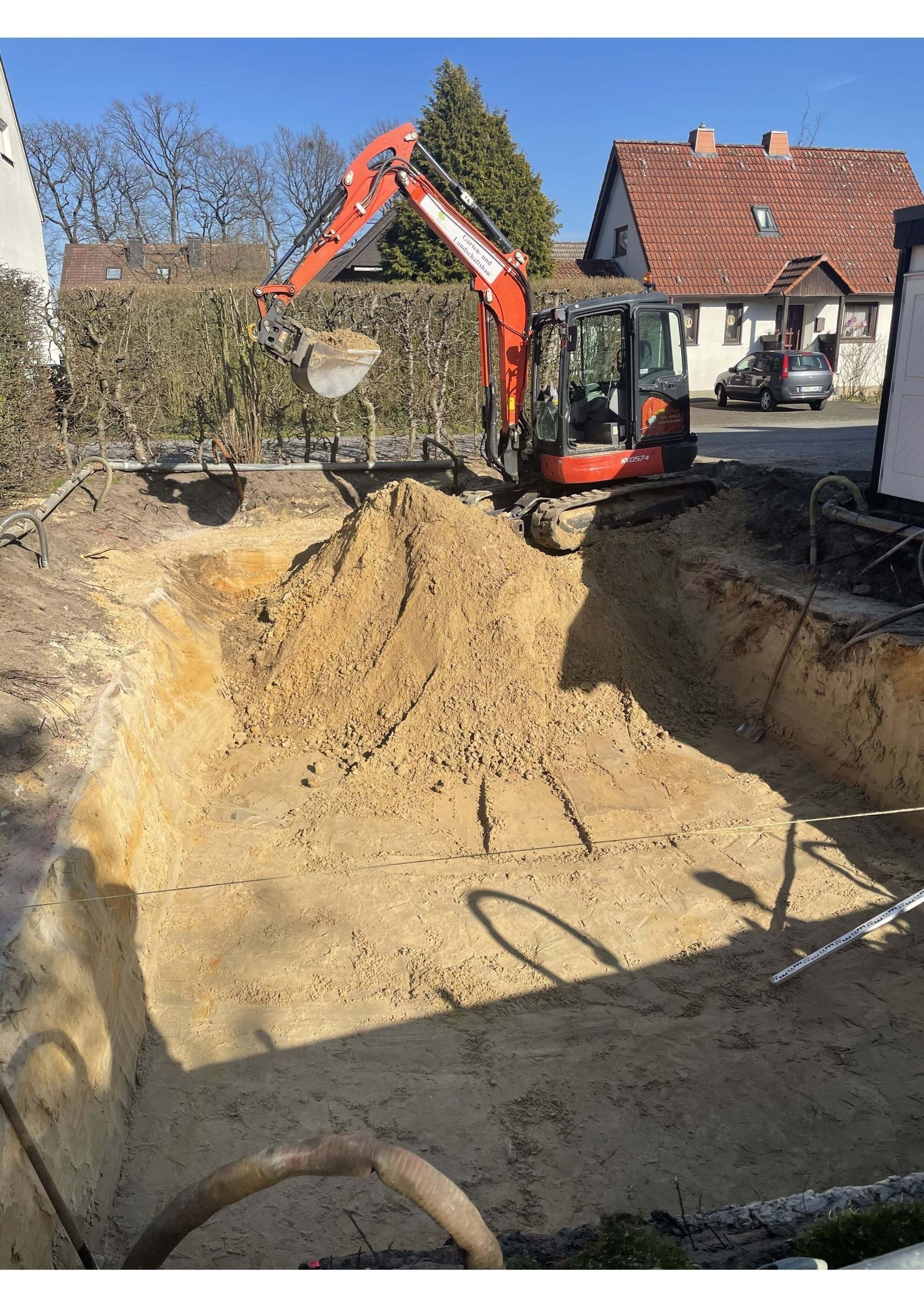 Ein Minibagger gräbt ein quadratisches Loch in einem Garten; in der Mitte ein Erdhaufen; im Hintergrund Häuser und eine Hecke.