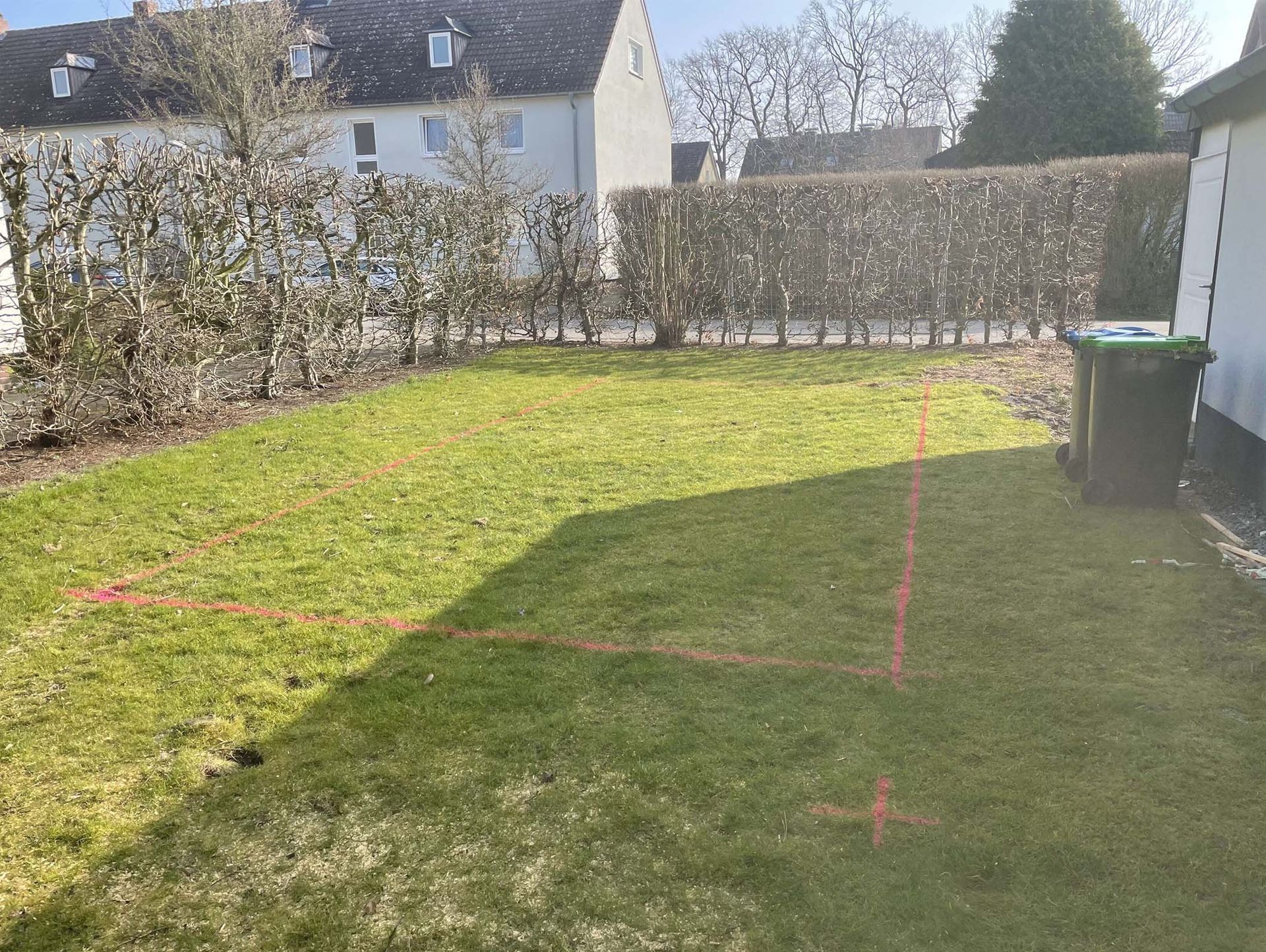 Grüner Rasen mit orangefarbenen Linien darauf, neben einer Hecke und Gebäuden unter blauem Himmel.