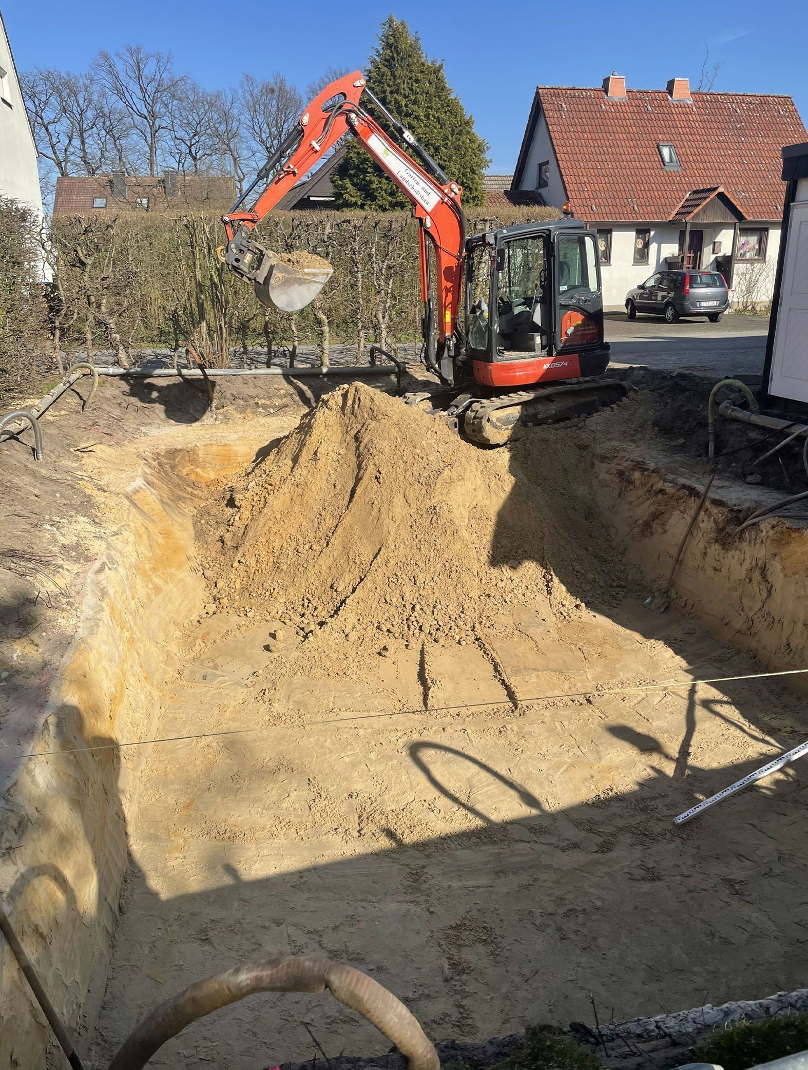 Ein Bagger gräbt ein rechteckiges Loch in einem Garten. Orangefarbene Maschine, braune Erde, sonniger Tag.