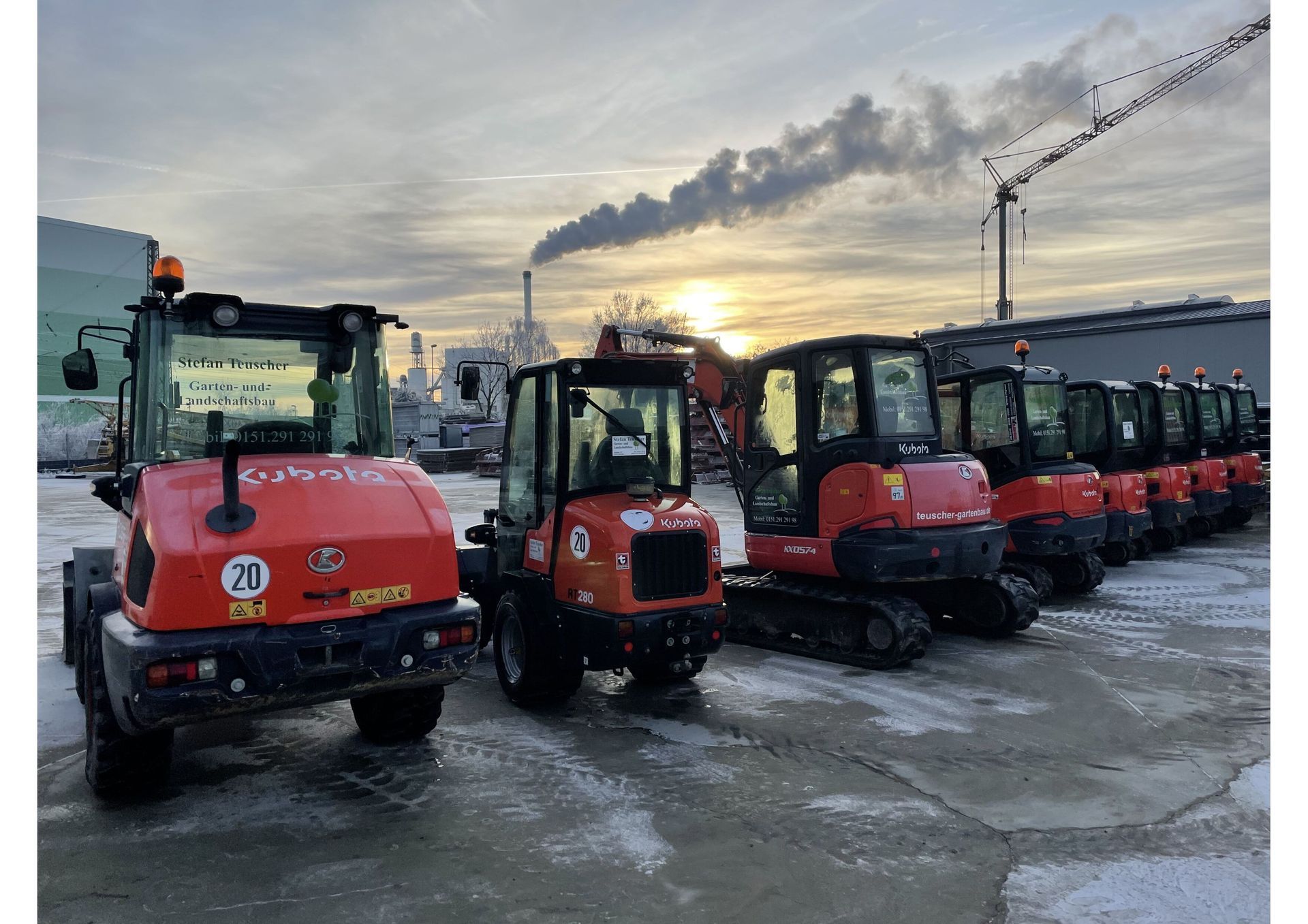 Eine Reihe orangefarbener Baufahrzeuge parkt auf dem Schnee, im Hintergrund ist eine Fabrik zu sehen.