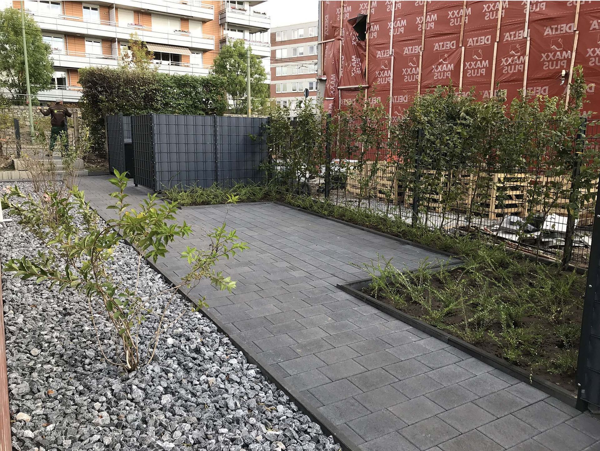 Ein gepflasterter Weg mit Landschaftsgestaltung, darunter Büsche, Kies und im Hintergrund ein im Bau befindliches Gebäude.