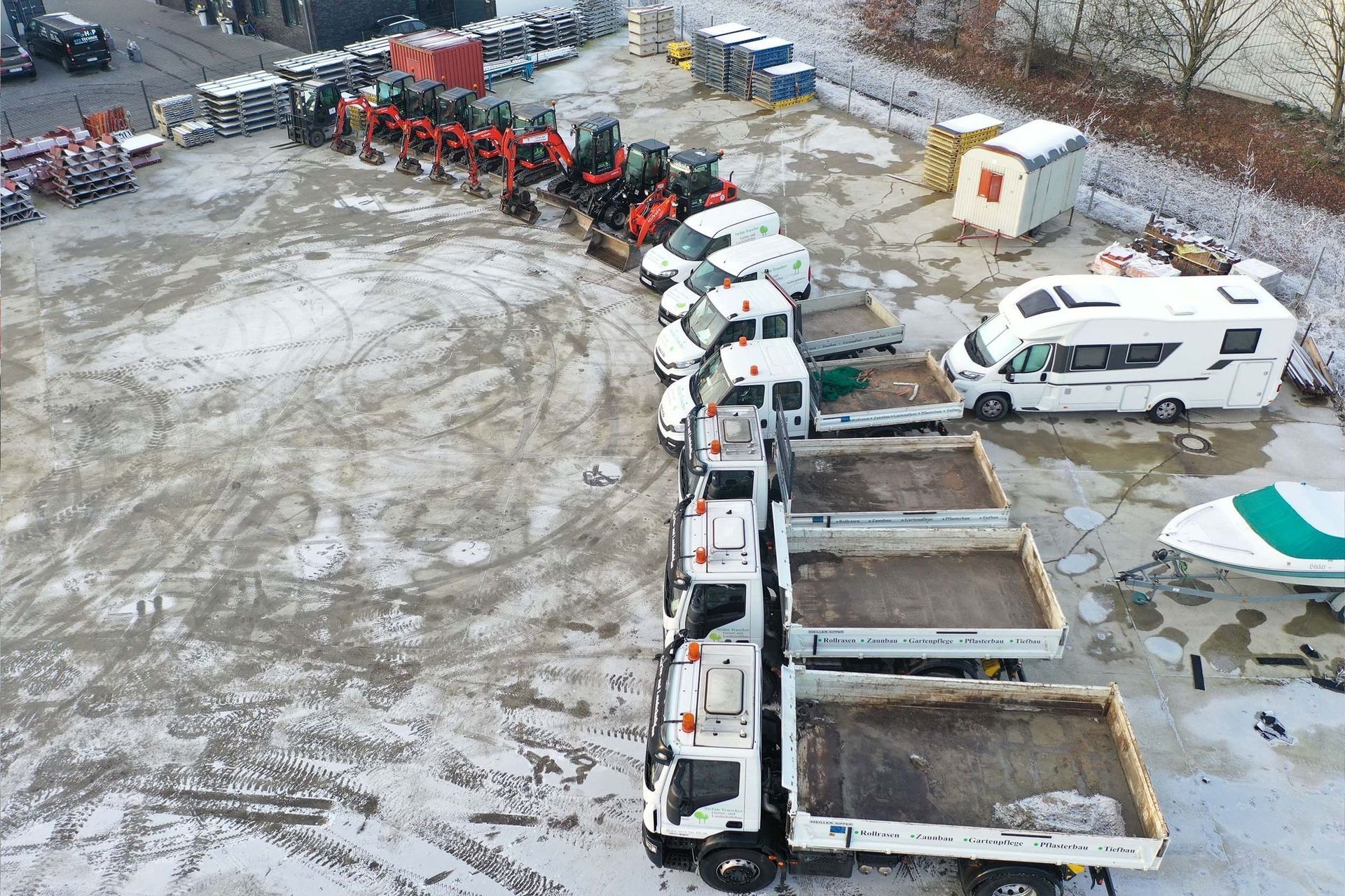Verschneiter Parkplatz mit geparkten Lastwagen, Traktoren und einem Wohnmobil.