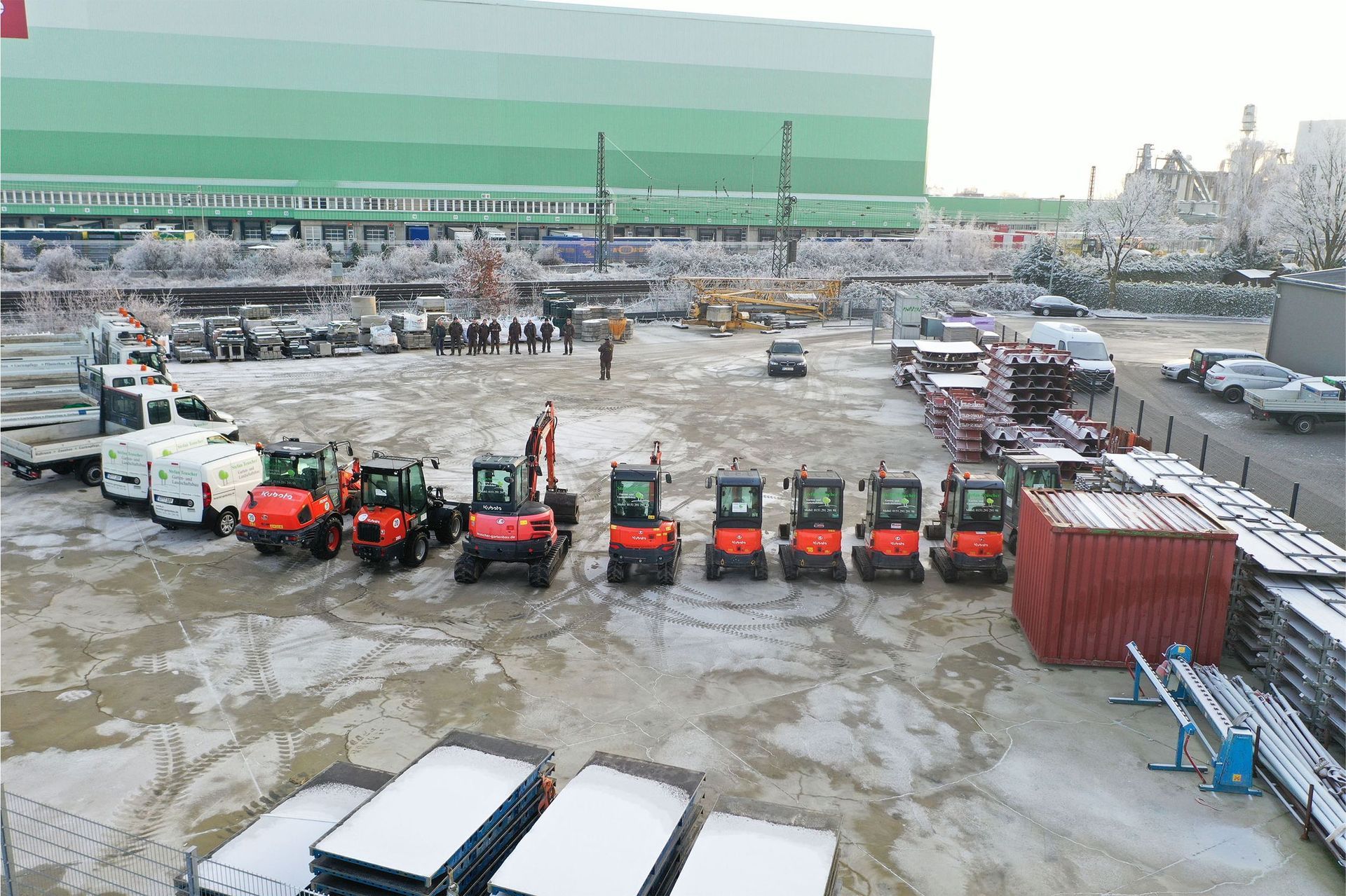 Eine Reihe orangefarbener Baustellenfahrzeuge parkt auf schneebedecktem Boden in der Nähe eines großen grünen Gebäudes.