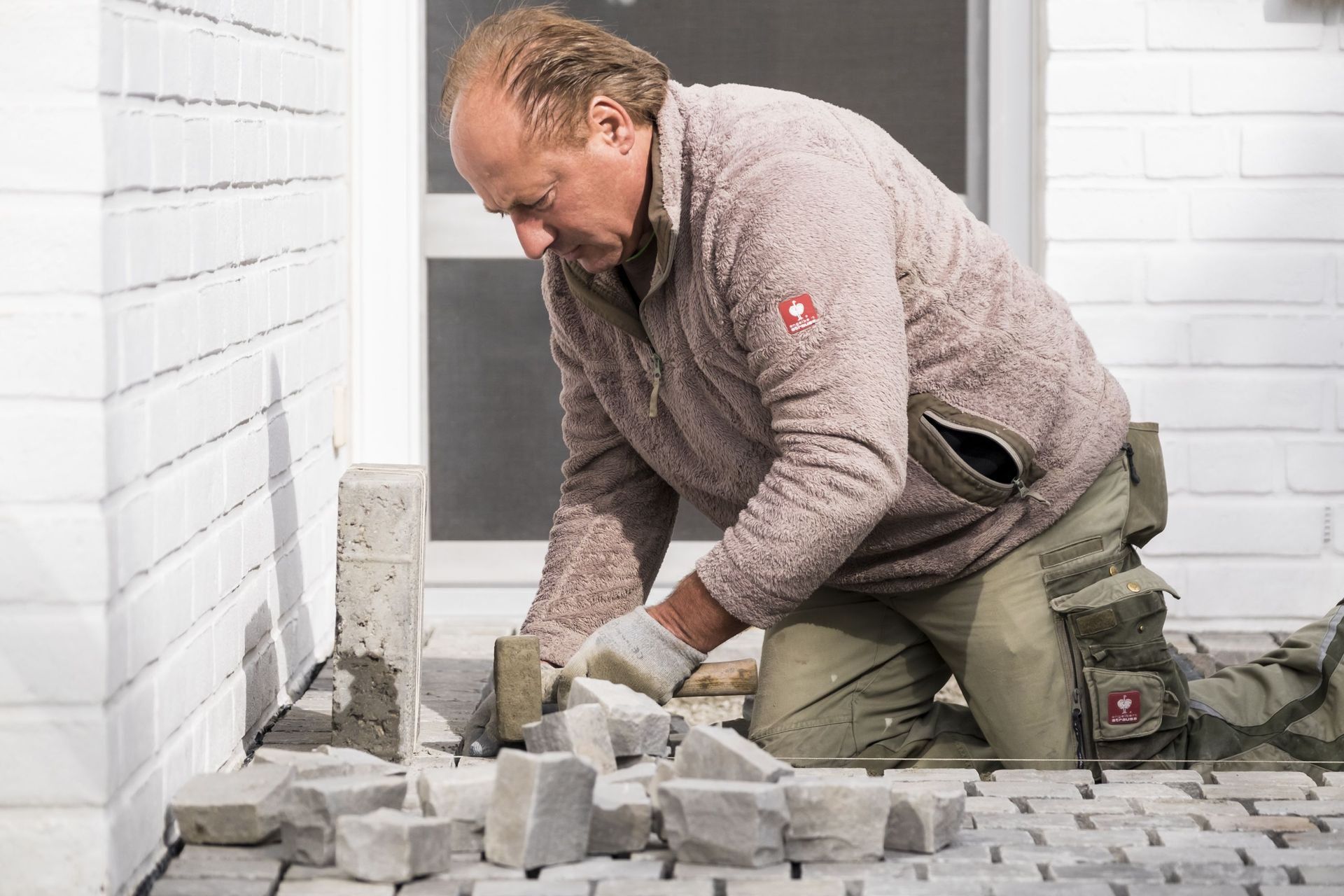 Ein Mann kniet und verlegt Pflastersteine ​​auf einer Terrasse neben einer weißen Backsteinmauer.