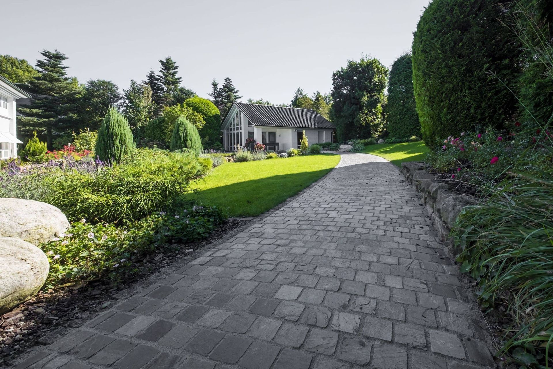 Ein Steinpfad führt zu einem weißen Haus mit grünem Rasen, umgeben von üppigen grünen Sträuchern und Bäumen.