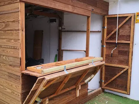 Un cobertizo de madera con la puerta abierta y una mesa de madera delante de él.