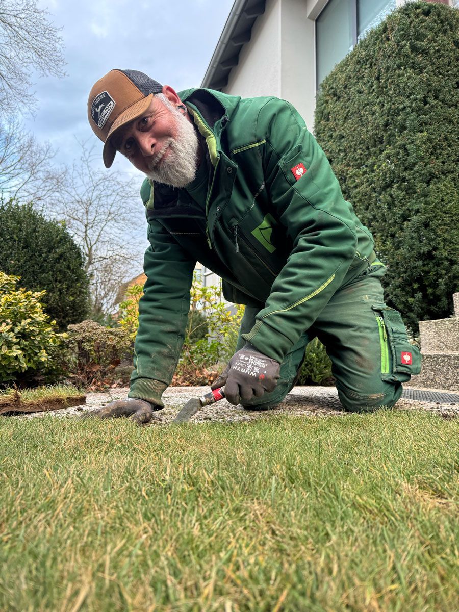 Ein Mann mit Bart kniet in einem Garten im Gras.