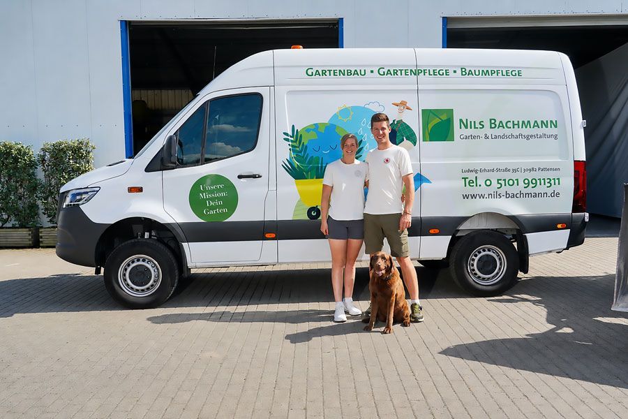 Zwei Personen mit einem Hund stehen neben einem weißen Transporter vor einem Lagerhaus.