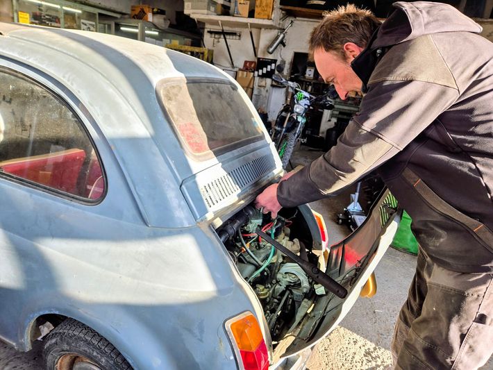 Une personne répare le moteur arrière d'une voiture ancienne bleu clair dans un garage.