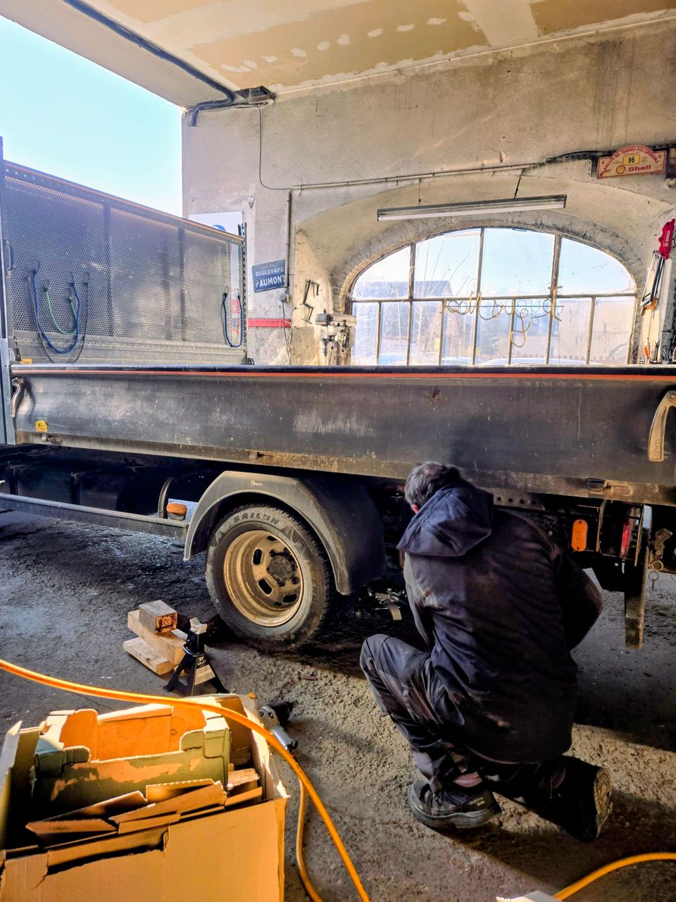 Un mécanicien est accroupi sous un camion plateau, travaillant sur la roue arrière dans un garage.