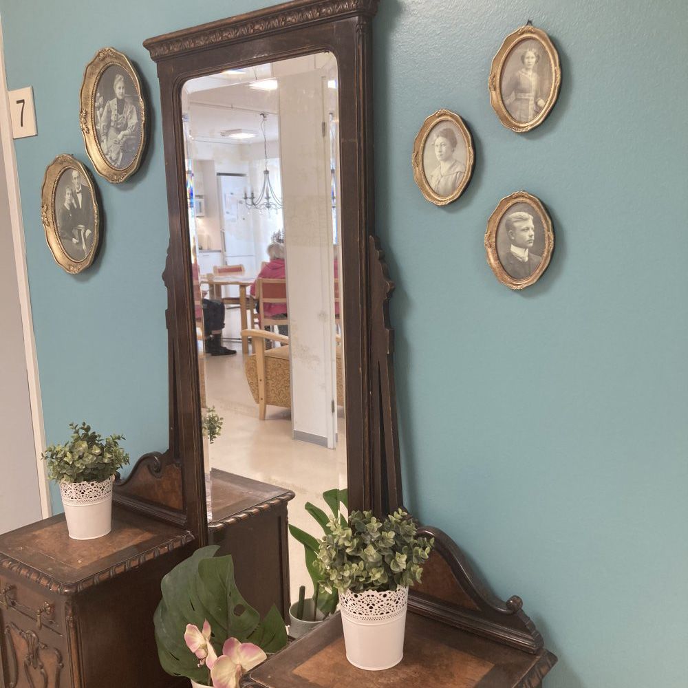 Antiikkipeili ja kasveilla koristeltuja sivupöytiä sinisellä seinällä, jolla on kehystettyjä muotokuvia.