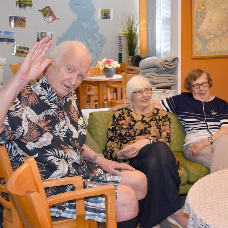 Kolme iäkästä ihmistä istuu yhdessä huoneessa. Yksi vilkuttaa hymyillen. Muut katsovat sivusta istuen sohvalla.