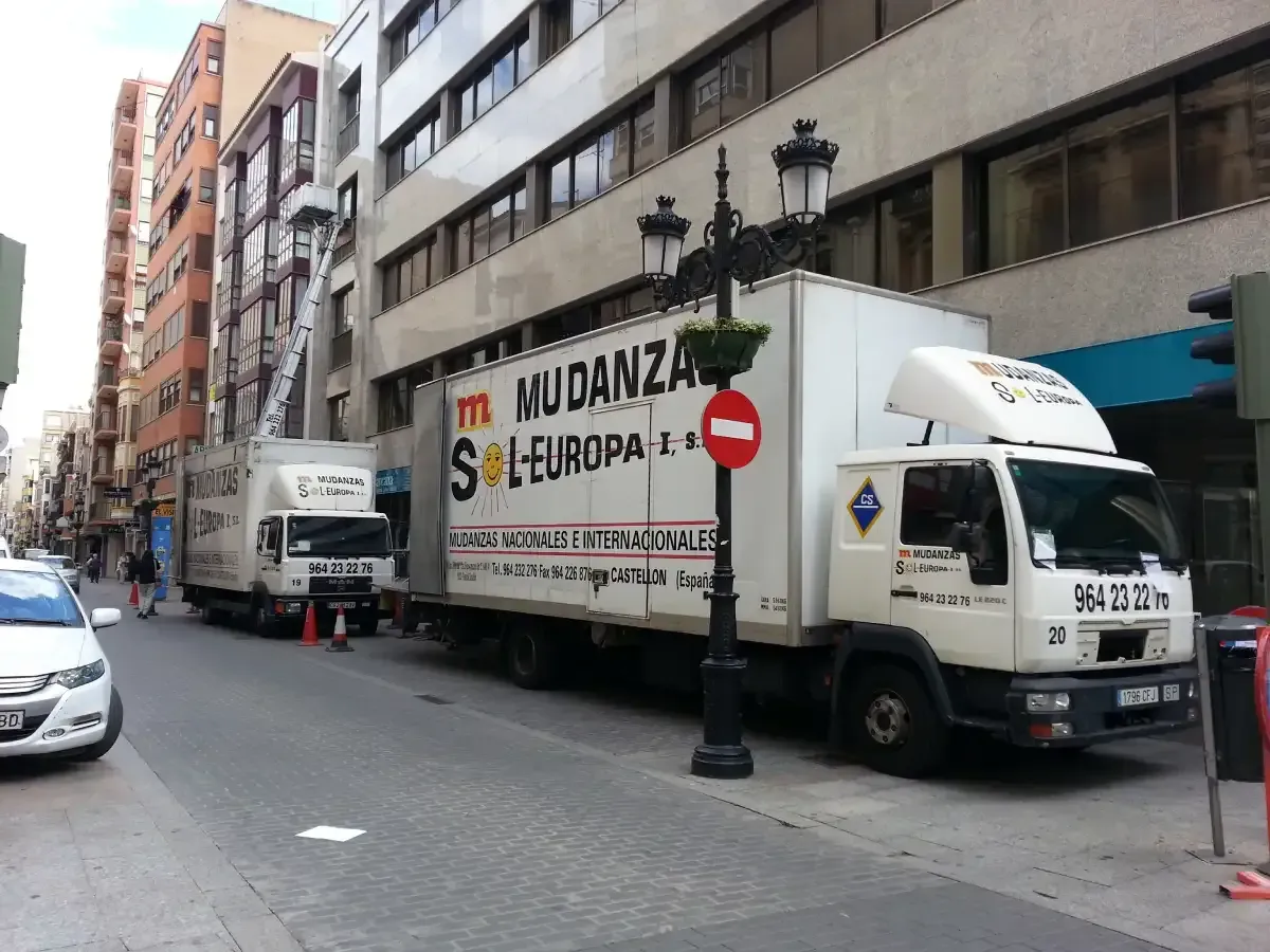 Dos camiones de mudanzas estacionados en una calle de la ciudad, cargando y descargando mercancías. Edificios a ambos lados.